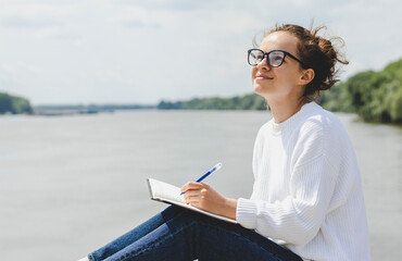 Young woman student in glasses writes entires in notebook while sitting outdoors. - obrazy, fototapety, plakaty