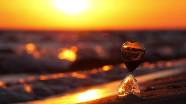 Small hourglass standing on beach against backdrop of bright sunset. The bright and rich warm color of sunlight reflects off water and wet sand. Wave run up and water wash ground around sand-glass