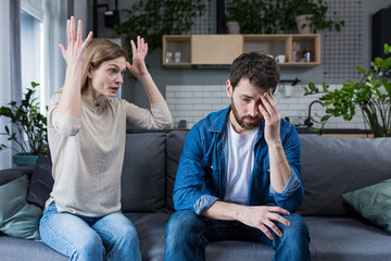 Family quarrel, man and woman sitting on sofa at home. angry woman yells at her husband, blames. The man is silent. He listens, holds his head, embarrassed