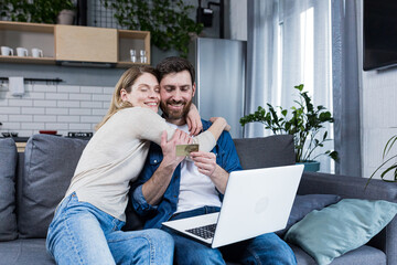 Online shopping. Man and woman shopping online from a notebook, holding a credit card in his hands. A purchase for a woman she chose herself. At home, sitting on the couch. Happy, satisfied