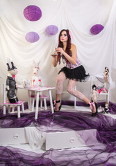 The girl, like a doll Alice in Wonderland, pours tea for dolls to rabbits from the tea set. Purple...