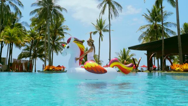 Excited young woman dancing and fun on floating toy in swimming pool in luxury tropical resort. Happy traveler woman in swimwear and hat relax on big inflatable unicorn pool float. Summer fun.