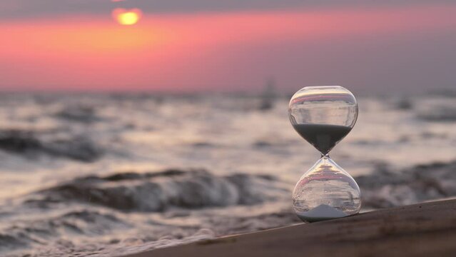 Sand-glass stand on sand against small waves of sea. Selective focus on foreground, blurred background. Continuous motion of waves, slow flow of sand grains in hourglass. Sun moves down and appears