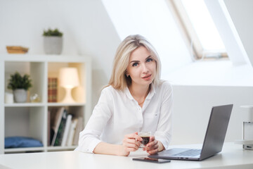 Young beautiful caucasian blonde woman working on laptop at home and drinking coffee. Remote work or freelance.