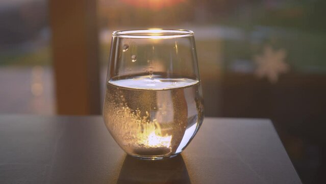 SLOW MOTION, CLOSE UP: Dissolving effervescent tablet dropped in a drinking glass backlit with the golden setting sun. Sparkling beverage for regaining health. Home treatment for seasonal illnesses.