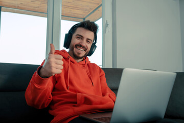 Young man listening to music with headphones making okay gesture with his hand, he has laptop in his lap and he is smiling