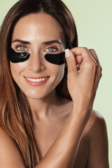 Fototapeta premium Portrait of smiling beauty woman, mature model, applies dark under eye patch from puffiness, anti-aging effect and facial skin lifting, has nourished glowing face, white background