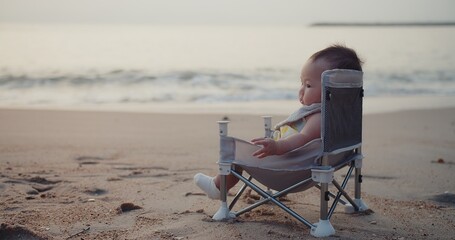 happy cute adorable Asian baby infant sitting relaxing on little chair with waves on background at seaside tropical sandy beach in sunrise during holiday vacation summertime Thailand	
