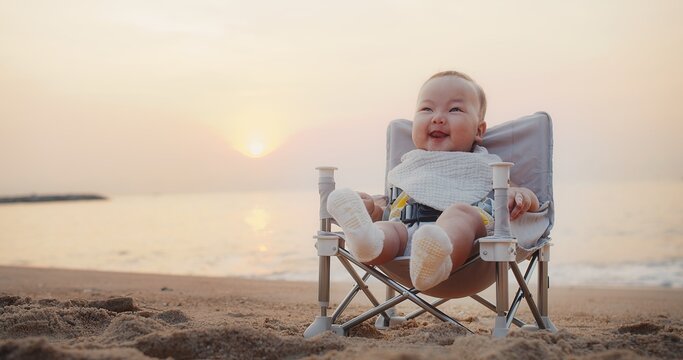 happy cute adorable Asian baby infant sitting relaxing on little chair and smiling with waves on background at seaside tropical sandy beach in sunrise during holiday vacation summertime Thailand	
