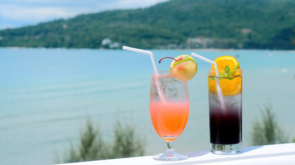 Two cocktail glasses with fruit decorations and a straw on on the railing of a luxury hotel resort with Andaman Sea on background. Fresh alcohol drinks and fruit cocktails.