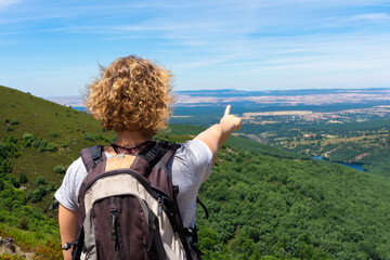 Blonde woman on her back with a backpack on her back, on top of a mountain, pointing with her hand...
