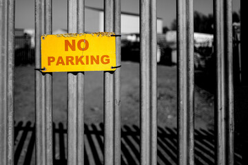 Shallow focus of a small No Parking sign seen on a metal gated entrance to an industrial yard. The sign is showing weathering around the edges.