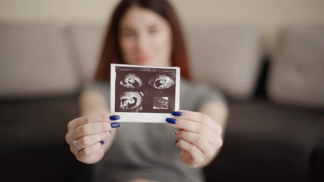 Pregnant woman in comfortable clothes sits on the floor and shows the camera an ultrasound photo of her baby