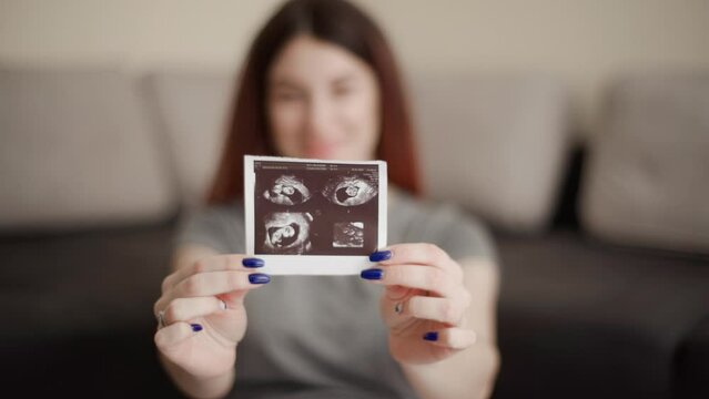 Pregnant woman in comfortable clothes sits on the floor and shows the camera an ultrasound photo of her baby