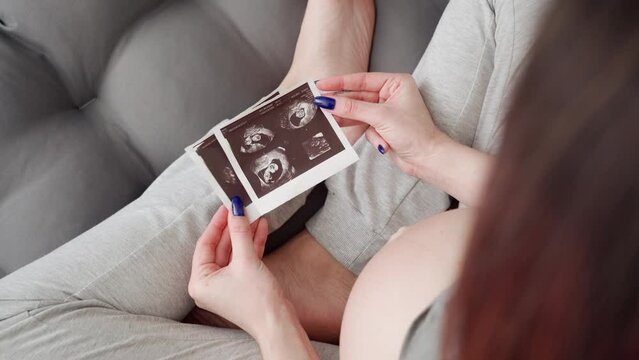 A pregnant woman in comfortable clothes sits on the floor and looks at an ultrasound photo of her baby.