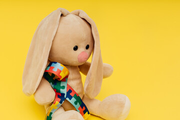 Toy bunny with puzzle pattern ribbon on a yellow background close-up. World autism awareness and pride day.