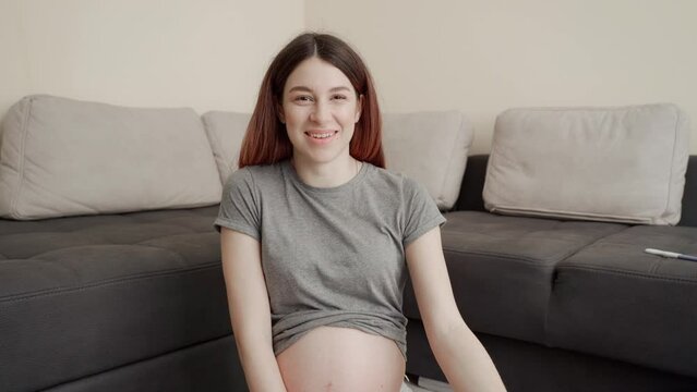 A young pregnant woman in a gray T-shirt sits on the floor, communicates with a friend and shows an ultrasound photo of her baby.