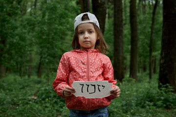 A little girl in a cap stands in a gloomy forest and holds a sign with the inscription in Hebrew...
