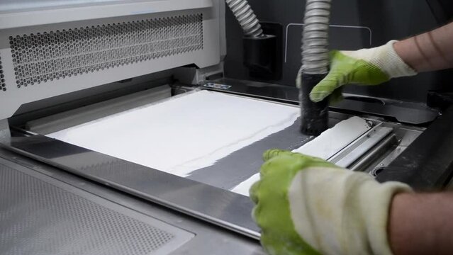 A male worker cleans the surface of an industrial 3D printer from white powder with a vacuum cleaner close-up. Multi Jet Fusion (MJF) 3D printing. The working process
