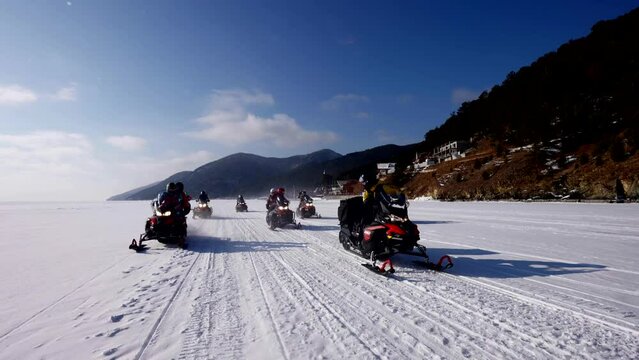 group of tourists are riding snow scooters on frozen lake or river in sunny winter day