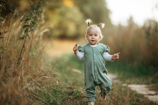 Cute Little Toddler Girl Learning To Walk In The Park, Adorable Kid Play With Grass. Plants, On A Warm Summer Day. The Concept Of Happy Childhood, Games. Family Leisure. Outdoor Play