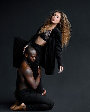 Front view of muscular tattooed afro american man, sitting on knees while supporting woman in black suit and laced bra which holding hand on his head and posing in black studio