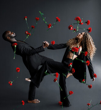 Side view of multiracial barefoot couple of dancer, man and woman closing eyes, wearing in stylish black suit and holding hands and leaning back while red flowers falling from above