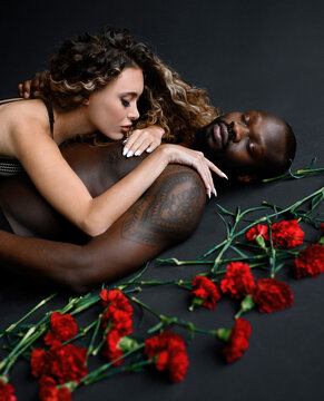 Cropped of passionate woman with Afro hairstyle, wearing in bra, lying on naked handsome tattooed Afro American man with closing eyes near scattered red carnations