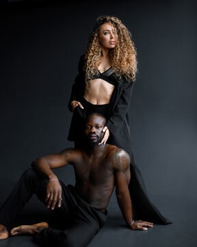 Front view of elegant curly haired woman in black oversize suit and bra, looking away while caressing face of muscular Afro American man in trousers which sitting on black background