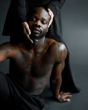 Cropped of calm Afro American male model with closed eyes and tattoos on muscular arms, sitting on floor in isolated studio while hands of faceless woman with white manicure touching his face