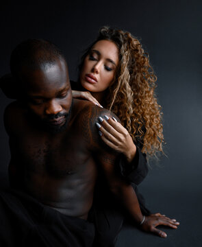 Cropped of loving couple, dark-skinned muscular man in trousers, sitting and looking down while beautiful woman with afro hairstyle and white manicure embracing behind back, posing indoors