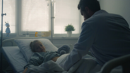 Pediatrician examining little patient in hospital ward. Girl lying in clinic bed