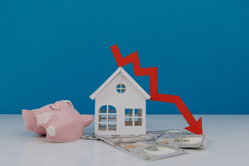 White model of house and broken piggy bank with down arrow. Falling of real estate market prices