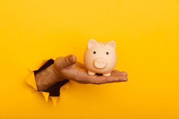 Hand holds a piggy bank through a hole in yellow paper wall