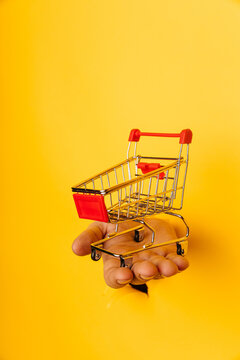 Male hand holds through a hole a mini grocery shopping trolley on a yellow paper background close-up. Vertical image