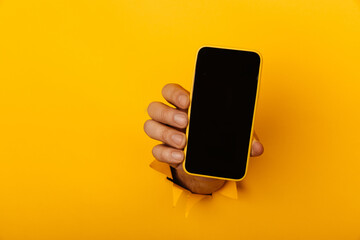Hand holds a smartphone out of a hole torn in yellow paper wall with copy space