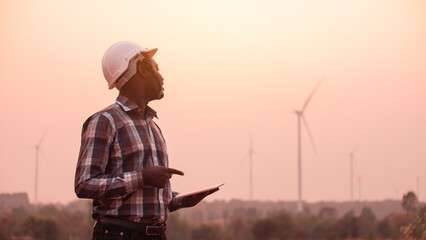 African engineer man stands holding tablet front the wind turbines generating electricity power...