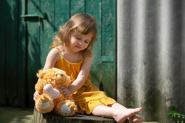 a little girl plays in the courtyard of the house on a sunny summer day with a toy soft bear