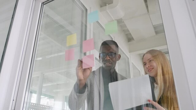 Two start-up business partners colleagues african man and caucasian young woman discuss their startup project, writing ideas on colorful stickers and put them on glass window