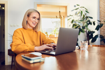 Portrait of middle aged woman sitting at dinning with laptop working at home - obrazy, fototapety, plakaty