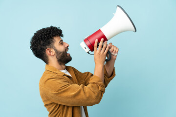 Young Moroccan man isolated on blue background shouting through a megaphone to announce something in lateral position