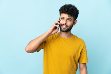 Young Moroccan man isolated on blue background keeping a conversation with the mobile phone with...