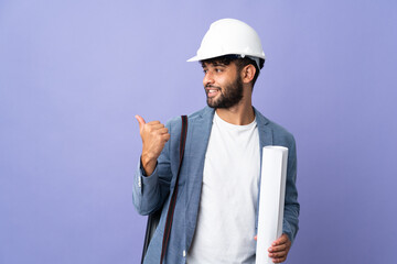 Young architect Moroccan man with helmet and holding blueprints over isolated background pointing...