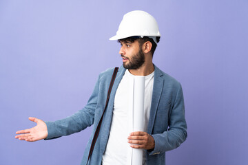 Young architect Moroccan man with helmet and holding blueprints over isolated background with...