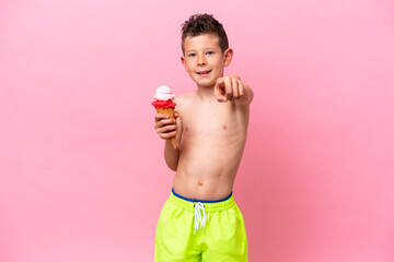 Little caucasian boy eating an ice-cream isolated on pink background points finger at you with a...