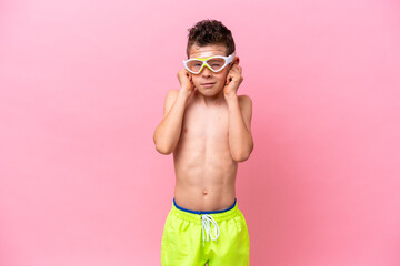 Little caucasian boy wearing a diving goggles isolated on pink background frustrated and covering ears