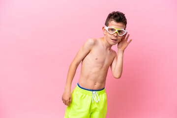 Little caucasian boy wearing a diving goggles isolated on pink background listening to something by...
