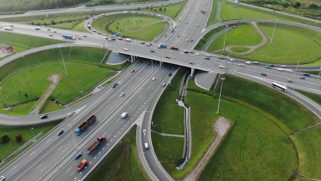 Cars drive along huge multi-level autobahn traffic intersection among green meadows against distant city in mist aerial panorama