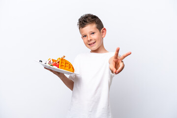 Little caucasian boy holding a waffles isolated on white background smiling and showing victory sign