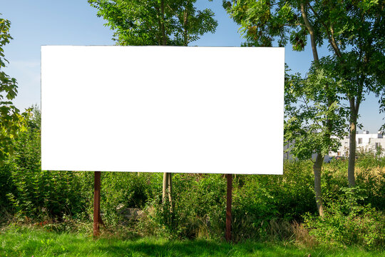 Blank white advertising billboard in fornt of green bushes. There are residential buildings in background.